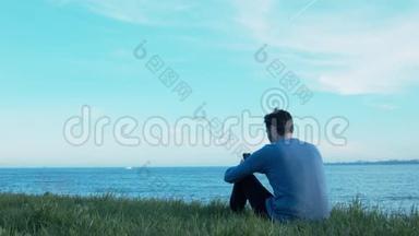 一个年轻人坐在海边的草地上写短信。 船在经过，飞机在空中飞行，